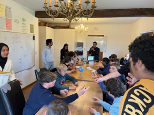 Un groupe d'enfants assis autour d'une table en train de participer à une activité, avec des adultes superviseurs. La scène se déroule dans une salle avec un tableau blanc et un lustre suspendu au plafond.