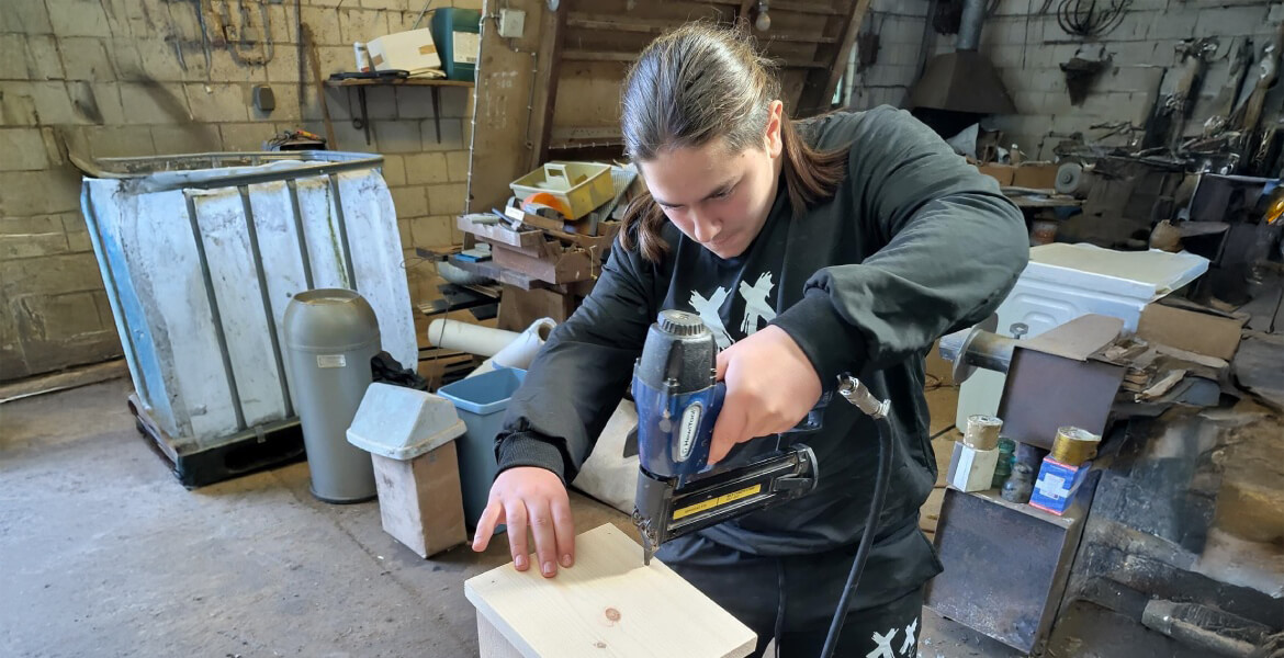 L'image montre une personne travaillant dans un atelier ou un espace de bricolage. La personne utilise une agrafeuse pneumatique pour fixer quelque chose sur un morceau de bois. L'atelier est rempli de divers outils et matériaux, y compris des bidons, des boîtes, des tuyaux et d'autres équipements. L'environnement semble être un espace de travail pratique, probablement utilisé pour des projets de construction ou de réparation.