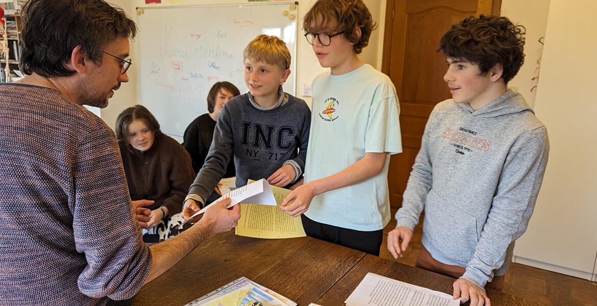 Dans cette image, un groupe de cinq personnes, dont quatre enfants et un adulte, est rassemblé autour d'une table. L'adulte, portant des lunettes et un pull à motifs, semble discuter ou donner des instructions aux enfants. Les enfants, debout autour de la table, écoutent attentivement et tiennent des feuilles de papier, probablement en train de participer à une activité éducative ou créative. L'un des enfants porte un t-shirt gris avec l'inscription "INC", tandis qu'un autre porte un t-shirt bleu clair avec un logo. À l'arrière-plan, on peut voir un tableau blanc avec des dessins et des notes, ce qui suggère que la scène se déroule dans un environnement d'apprentissage ou de travail collaboratif.