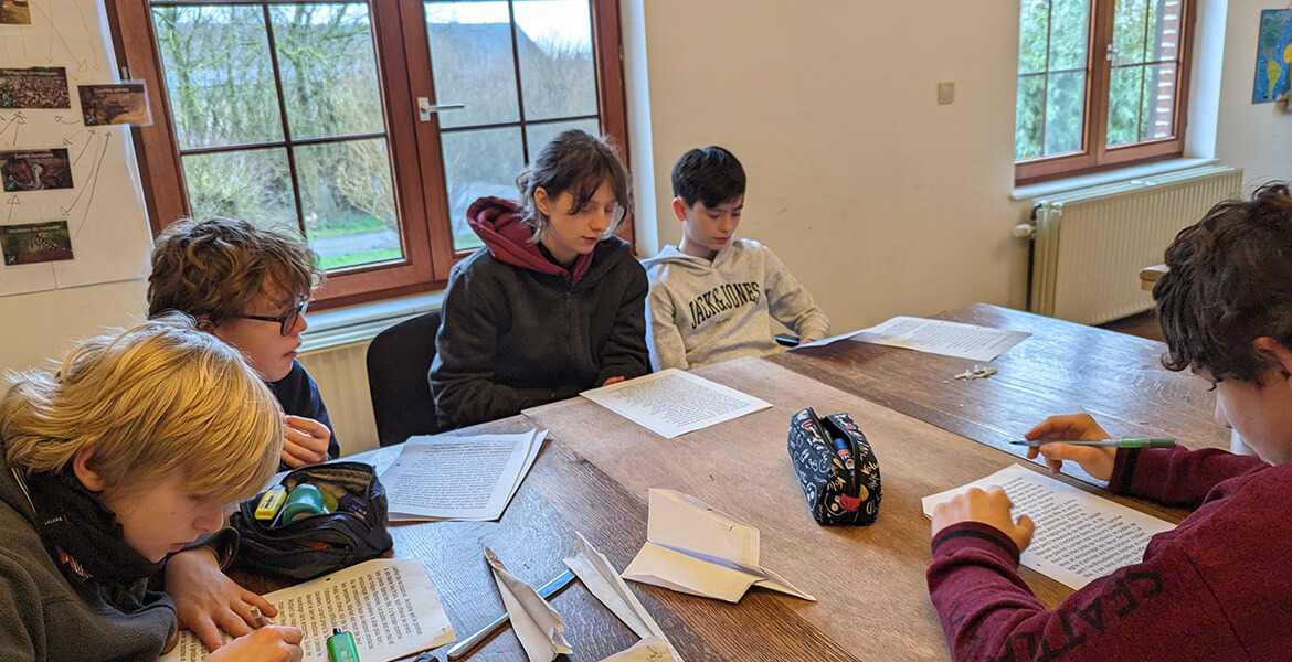 L'image montre un groupe de cinq enfants assis autour d'une table en bois dans une salle de classe. Ils sont tous concentrés sur des activités impliquant des feuilles de papier, probablement en train de lire ou d'écrire. La pièce est bien éclairée grâce à de grandes fenêtres laissant entrer la lumière naturelle. Des affiches et des dessins sont accrochés aux murs, et on peut voir des arbres à l'extérieur des fenêtres, indiquant un environnement calme et verdoyant. Un sac à crayons est posé sur la table, suggérant que les enfants sont en train de travailler sur un projet scolaire ou une activité créative.