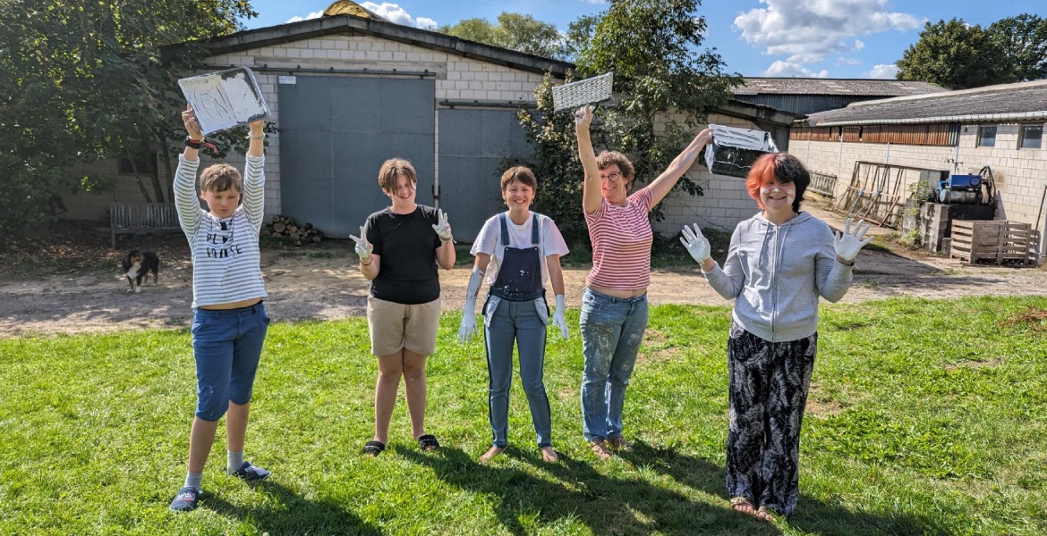 Cette image montre cinq personnes posant joyeusement dans un cadre extérieur, probablement une ferme ou un jardin. Elles sont debout sur une pelouse et tiennent des plateaux blancs au-dessus de leurs têtes. Les personnes portent des vêtements décontractés et certains d'entre eux ont des gants, ce qui suggère qu'ils pourraient être en train de participer à une activité pratique ou de jardinage. À l'arrière-plan, on peut voir des bâtiments en brique et un chien qui se promène, ajoutant à l'ambiance rurale et détendue de la scène. Le ciel est dégagé et ensoleillé, indiquant une belle journée.