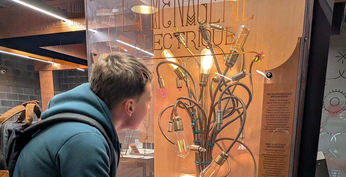 Un jeune homme observe une vitrine d'exposition intitulée 'Ménagerie Électrique', présentant une sculpture lumineuse composée de plusieurs ampoules et câbles entrelacés.