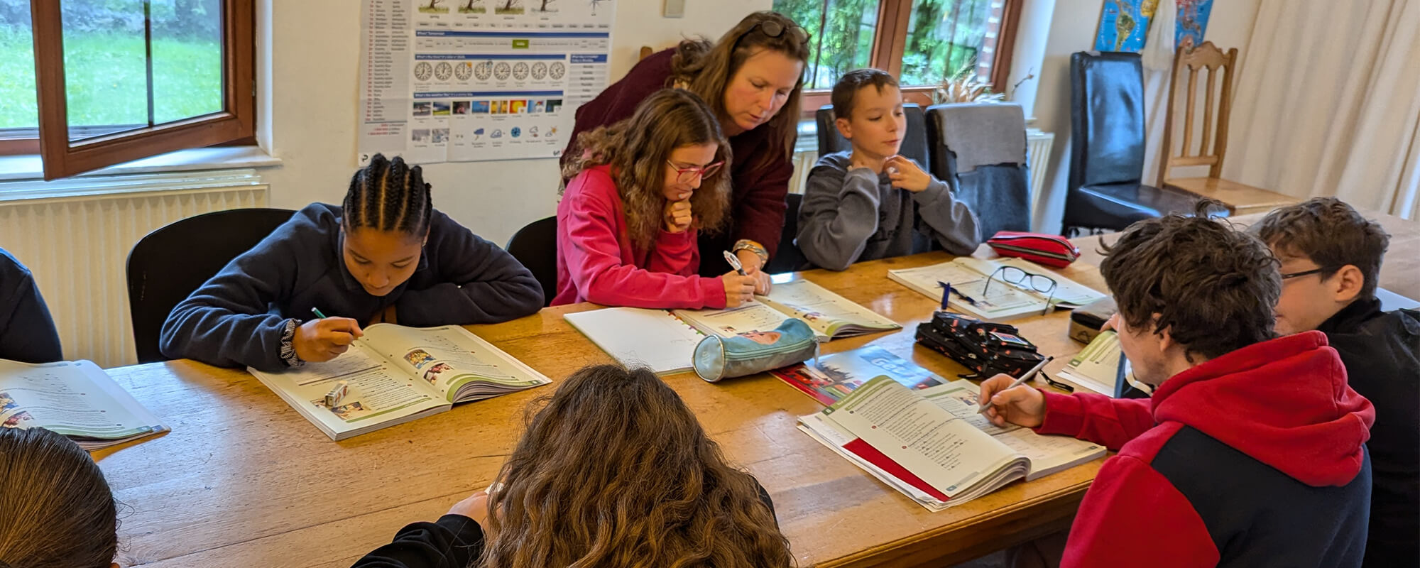 Un groupe d'enfants assis autour d'une table en train de travailler dans des cahiers, accompagnés par deux adultes qui les aident. La scène se déroule dans une salle de classe bien éclairée avec des fenêtres donnant sur un espace vert extérieur.