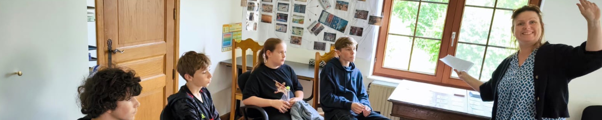 L'image montre un groupe de jeunes assis en cercle dans une salle de classe, participant à une activité ou une discussion. Une femme debout semble animer la session, tenant un morceau de papier et gesticulant, probablement pour expliquer ou poser une ques