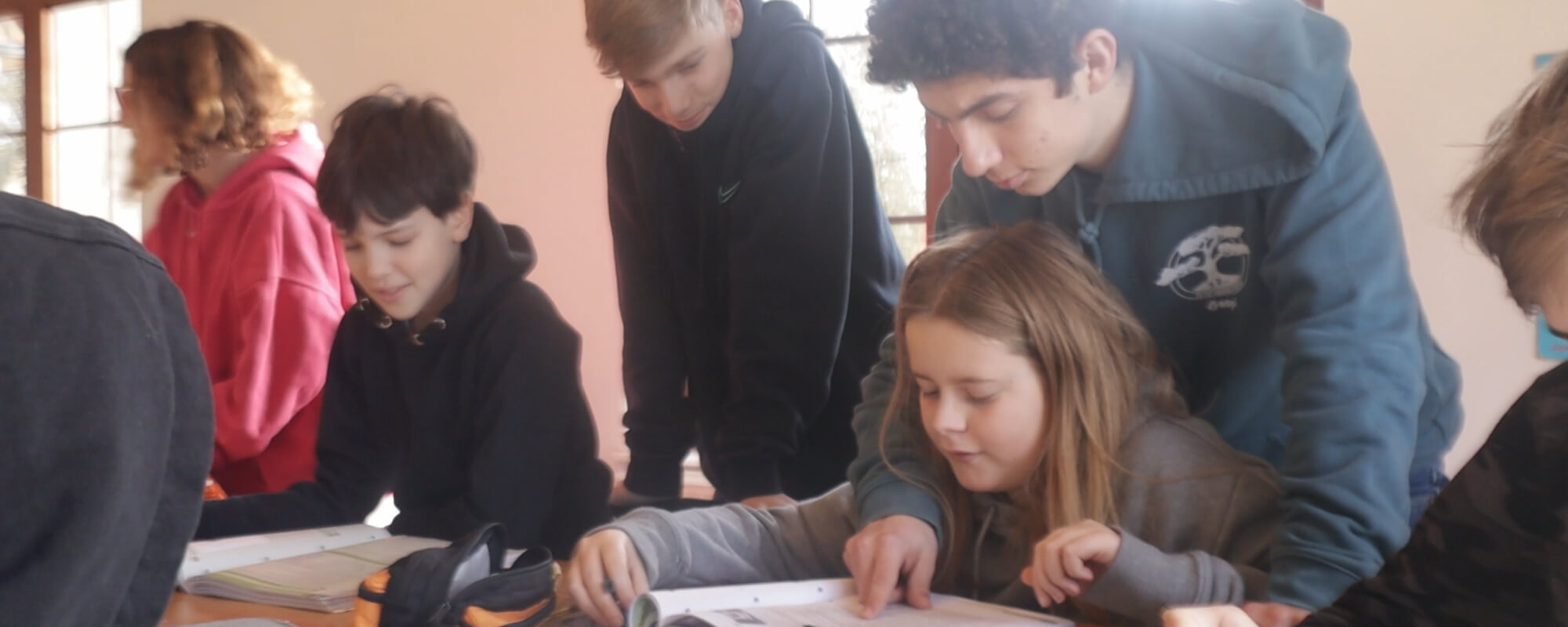 L'image montre un groupe de jeunes, probablement des élèves, rassemblés autour d'une table. Ils semblent être engagés dans une activité collaborative, peut-être un projet scolaire ou une tâche de groupe. Les élèves sont concentrés sur des documents ou des