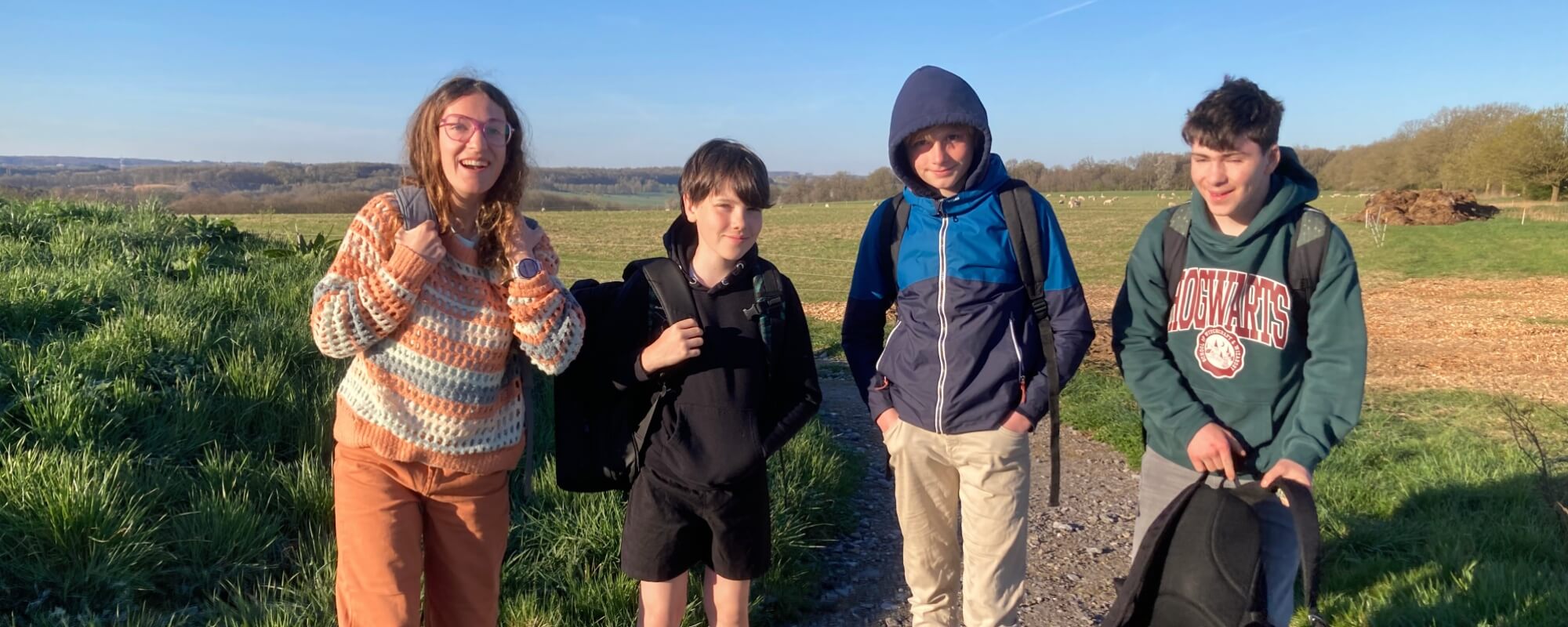 L'image montre quatre jeunes personnes posant joyeusement sur un chemin de terre en pleine nature. Ils sont vêtus de vêtements décontractés et portent des sacs à dos, suggérant qu'ils pourraient être en randonnée ou en excursion. Le paysage autour d'eux e