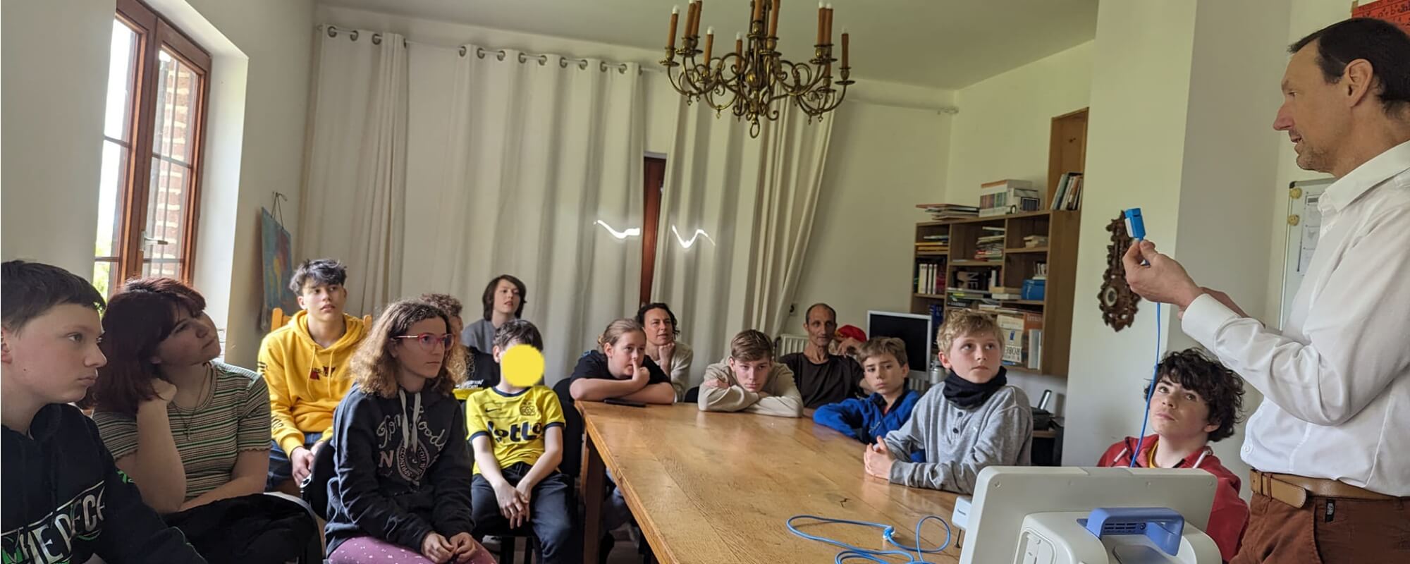 L'image montre un groupe d'enfants et d'adultes assis autour d'une table dans une salle de classe ou une salle de réunion. Ils semblent attentifs et engagés dans une activité ou une démonstration. Un homme à droite de l'image, probablement un enseignant o