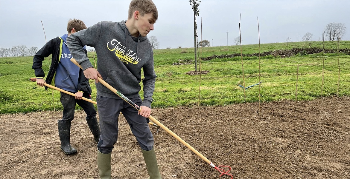 L'image montre deux jeunes hommes travaillant dans un champ. Ils sont équipés de bottes en caoutchouc et utilisent des outils manuels, probablement pour désherber ou préparer le sol. L'un d'eux porte un sweat-shirt gris avec une inscription et l'autre a u
