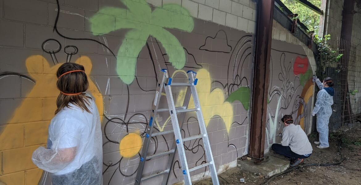 L'image montre trois personnes en train de peindre une fresque murale sur un mur en brique. Elles sont équipées de combinaisons de protection et utilisent des bombes de peinture en aérosol pour créer leur œuvre. Une échelle est placée contre le mur, perme
