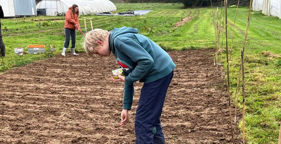 L'image montre deux personnes travaillant dans un jardin ou une ferme. La personne au premier plan, probablement un enfant, est penchée et semble planter ou examiner quelque chose dans le sol. Elle tient un petit objet dans sa main droite. La personne à l