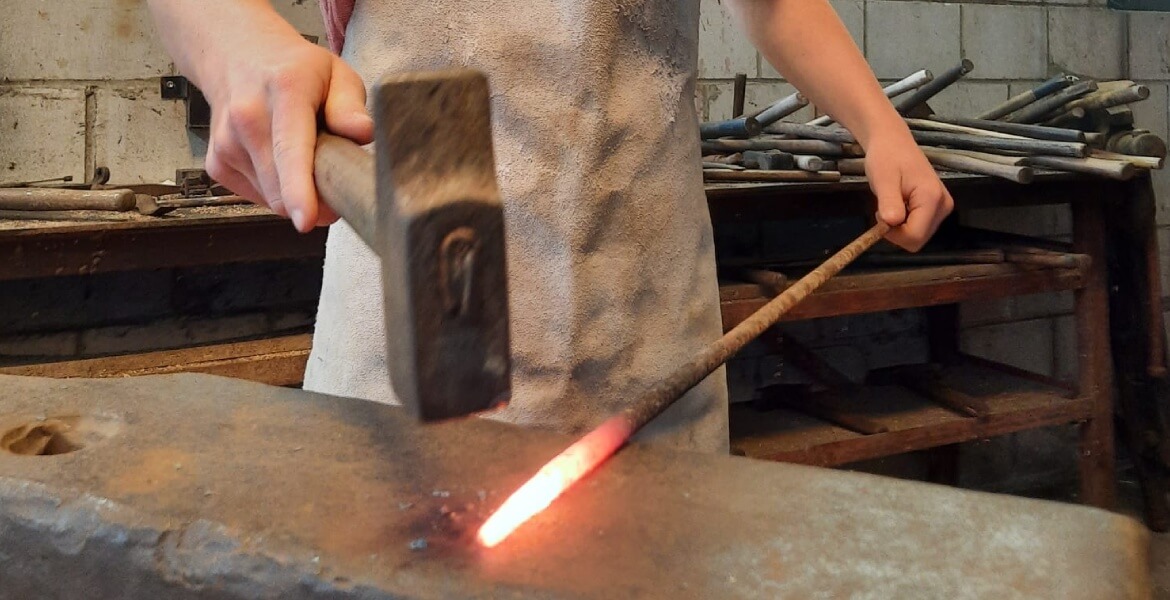 L'image montre une personne en train de travailler dans un atelier de forge. La personne utilise un marteau pour frapper un morceau de métal chauffé au rouge, posé sur une enclume. Elle porte un tablier de protection et manipule le métal avec des pinces. 