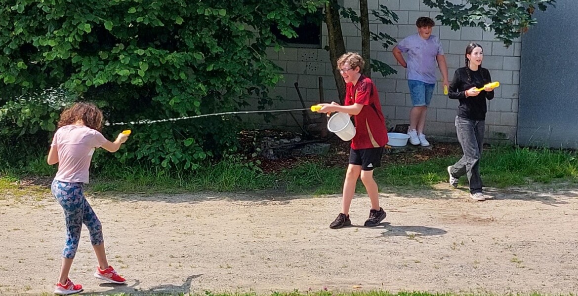 L'image montre un groupe de jeunes en train de jouer avec des pistolets à eau dans un espace extérieur. L'un des jeunes, un garçon en rouge, tient un seau et semble être en train de remplir ou de préparer un pistolet à eau. Une fille en premier plan est e