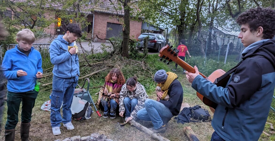 L'image montre un groupe de jeunes personnes réunies autour d'un feu de camp en plein air. Certains sont assis sur des bûches ou debout, et ils semblent profiter d'un moment de détente et de camaraderie. L'un des jeunes joue de la guitare, ajoutant une am