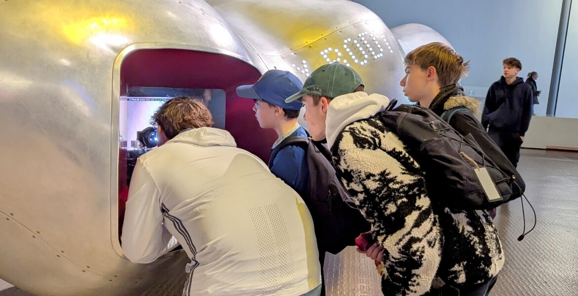 L'image montre un groupe de jeunes personnes dans ce qui semble être un espace d'exposition ou un musée. Ils sont regroupés autour d'une fenêtre ou d'une ouverture dans une structure métallique, probablement en train d'observer quelque chose à l'intérieur