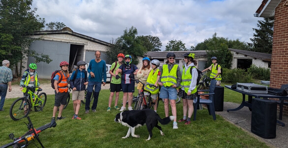 L'image montre un groupe de jeunes et d'adultes posant ensemble à l'extérieur, probablement avant ou après une activité de vélo. Ils sont équipés de casques et de gilets fluorescents, indiquant une attention particulière à la sécurité. Plusieurs vélos son