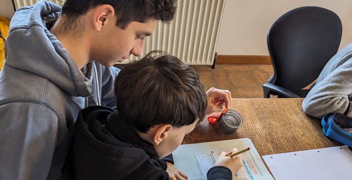 L'image montre deux jeunes personnes assises à une table, engagées dans une activité d'écriture ou de dessin. L'un d'eux, probablement un adolescent, semble aider ou superviser l'autre, un enfant plus jeune. L'enfant tient un crayon et travaille sur une f