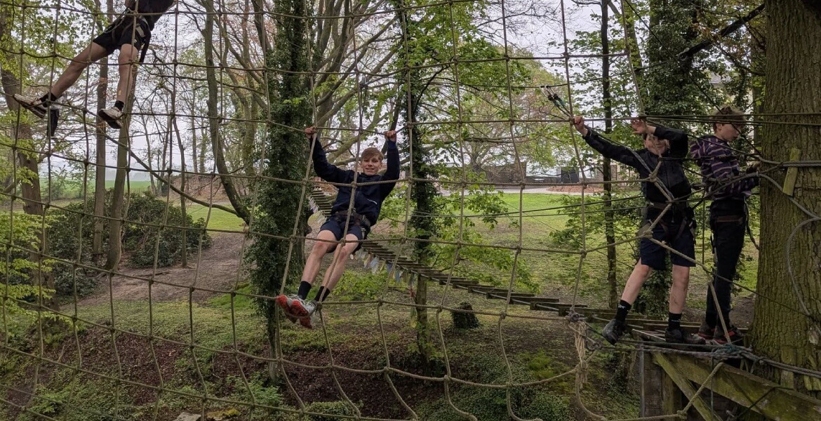 L'image montre un groupe de jeunes en train de participer à une activité de parcours aventure en forêt. Ils sont équipés de harnais de sécurité et évoluent sur un parcours d'escalade composé de filets et de cordes entre les arbres. Les jeunes semblent con