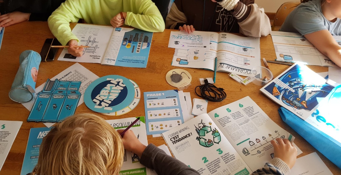 L'image montre un groupe d'enfants assis autour d'une table, engagés dans une activité éducative. La table est couverte de divers documents et outils pédagogiques, y compris des livrets, des cartes, des crayons, et des jeux de société éducatifs. L'un des 