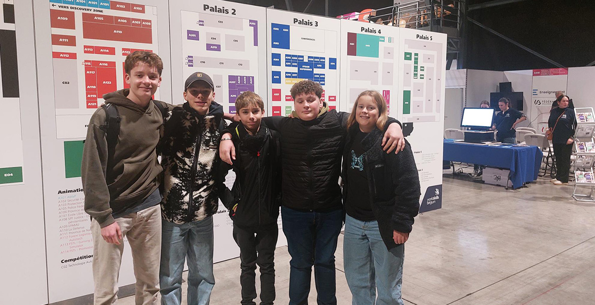 L'image montre un groupe de quatre jeunes personnes posant ensemble devant des panneaux d'affichage dans ce qui semble être un salon professionnel ou une foire. Les panneaux derrière eux sont intitulés 