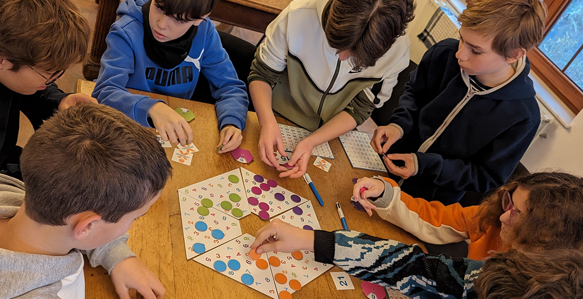 Dans la deuxième image, un autre groupe d'enfants est rassemblé autour d'une table, jouant à un jeu de société qui implique des cartes et des plateaux avec des cercles colorés et numérotés. Les enfants semblent concentrés et impliqués dans le jeu, interag