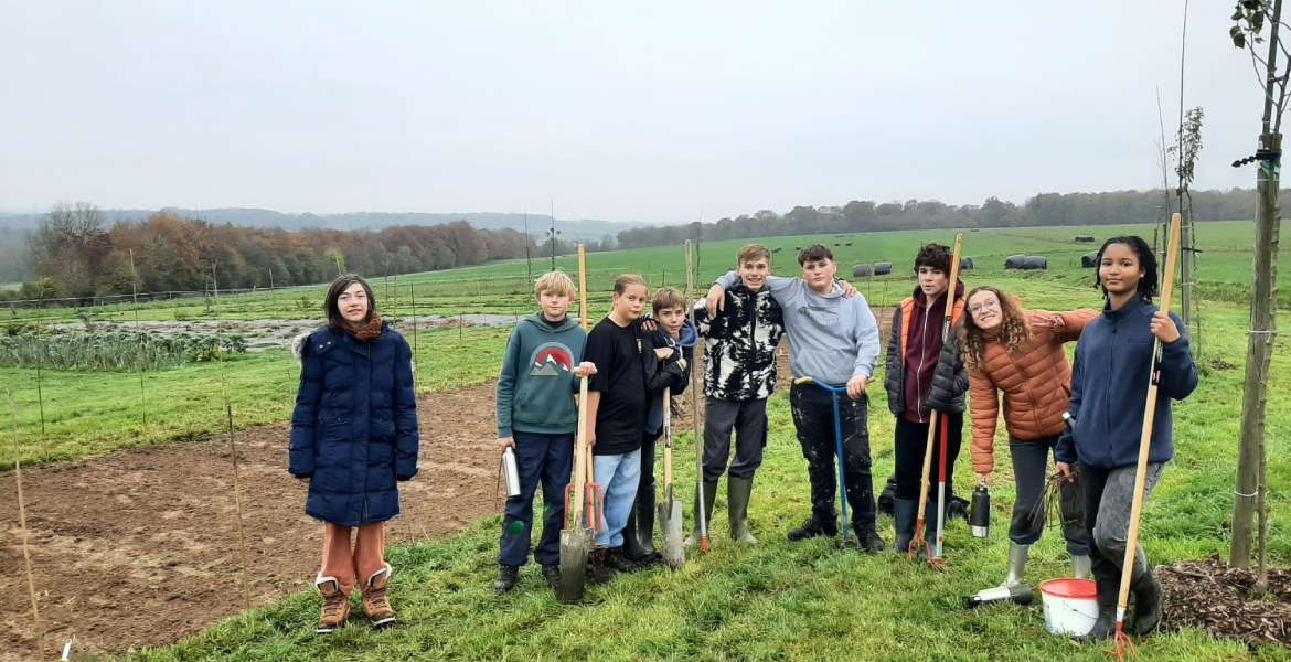 L'image montre un groupe de jeunes personnes posant ensemble dans un champ ou un jardin. Ils sont équipés d'outils de jardinage tels que des pelles, des râteaux et des fourches, ce qui suggère qu'ils participent à une activité de jardinage ou de plantatio